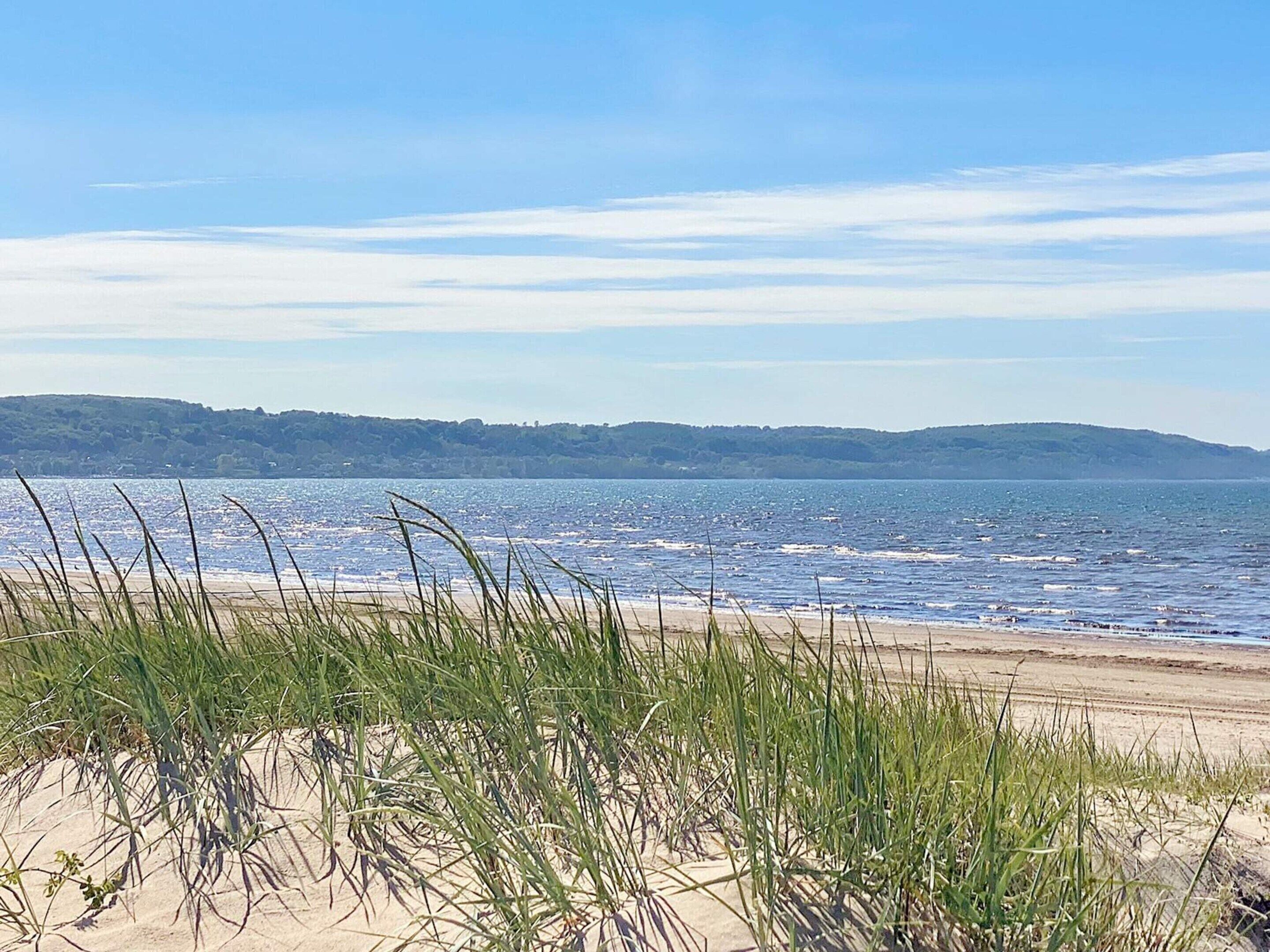 Une plage avec des dunes de sable et de l’herbe au premier plan, une mer calme et un paysage vallonné à l’horizon, sous un ciel dégagé.