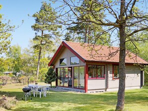 Une petite maison à un seul étage, avec un toit rouge et des murs blancs, entourée d’arbres et d’une pelouse verdoyante.