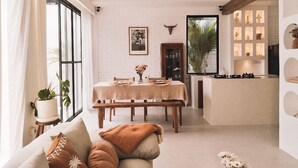 A dining area with a table set for four, a sofa with cushions, a potted plant, and a kitchen area with a stove and cabinets.