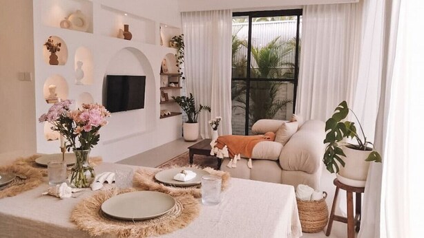 A modern living room with a sofa, a dining table set with plates and a vase of flowers, and a large window with curtains.