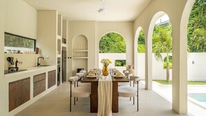 Een moderne eetruimte met een lange tafel voor zes personen, omringd door stoelen. Er is een plafondventilator, een keuken met kasten en een aanrecht, en uitzicht op een zwembad door bogen.