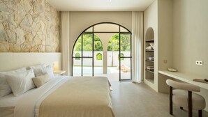 Een slaapkamer met een groot bed, een stenen muur, een bureau met stoel, en uitzicht op een tuin door een glazen deur.