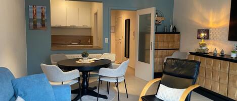 A modern living room with a round dining table, light grey chairs, a blue sofa, and a black leather chair. There is a kitchen area with wooden cabinets and a built-in oven.