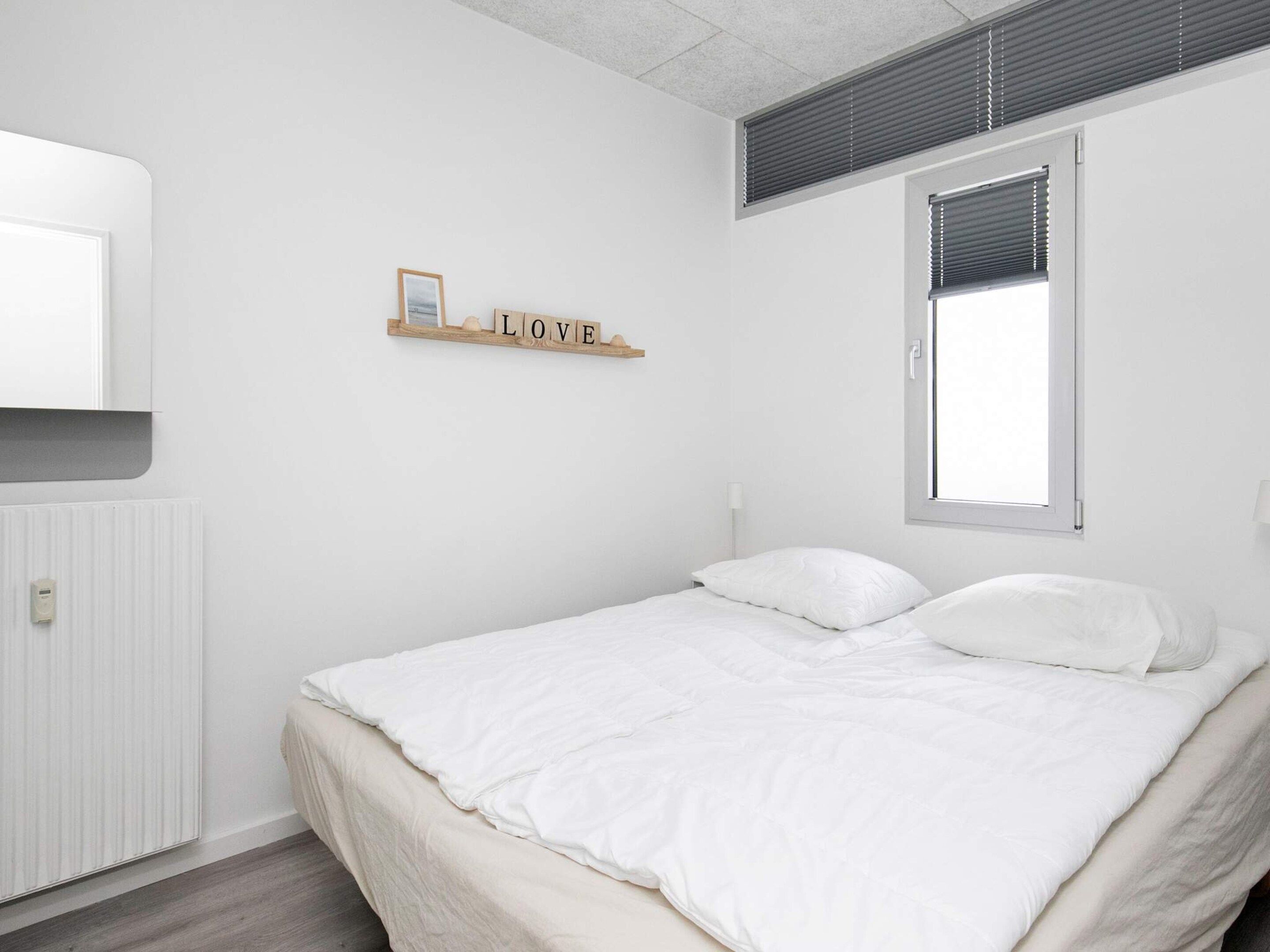 A bedroom with a bed, a radiator, a window with blinds, and a wall-mounted shelf with the word "LOVE".