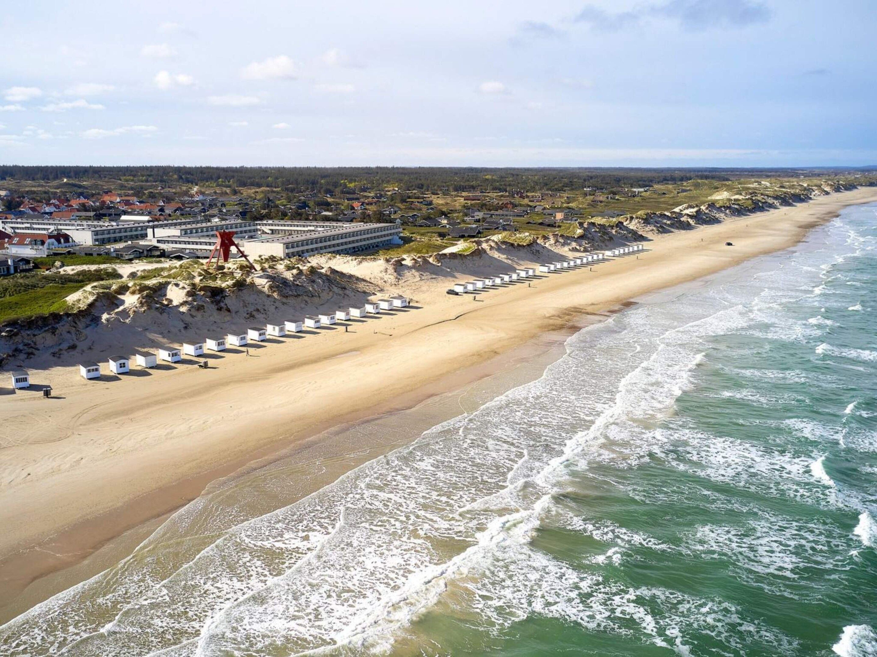 Ein Strand mit einer Reihe weißer Strandhütten, ein großes Gebäude mit zahlreichen Fenstern und ein sandiger Uferstreifen mit Wellen.