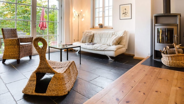 A cozy living room with a sofa, wicker chairs, a wooden table, a fireplace, and large windows with a view of greenery. - Holiday Apartment Remise - Ground Floor (Werder (Havel))