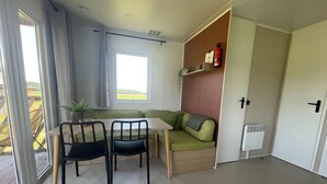Una habitación pequeña con mesa de comedor, dos sillas, un sofá verde, una ventana con cortinas y un radiador.