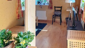 Una habitación acogedora con suelo de madera, ventana con cortinas, radiador, una silla, una mesa con lámpara y plantas en macetas.
