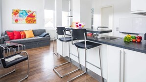 A modern kitchen with white cabinets, a black countertop, and stainless steel appliances. There are two bar stools and a dining table with chairs. A colorful abstract painting is on the wall, and a sofa with red cushions is in the living area. - Lively Apartment in Norderney (Norderney)