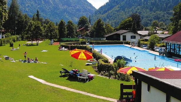 A pool area with lounge chairs and umbrellas, surrounded by mountains and greenery. - Ferienwohnung in Bayrischzell (Bayrischzell)