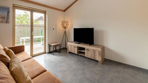 Sala de estar com sofá, TV sobre suporte de madeira, luminária de chão e porta de vidro que se abre para o exterior.