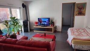A living room with a red sofa, a TV on a wooden stand, and a bed with a quilt. There are potted plants and a red lamp on the stand. - Cottage alsace bossue (Weyer)