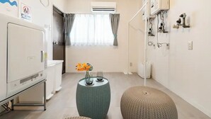 A compact hotel room with a kitchenette, a small round table with a vase of flowers, and a round ottoman. - Wagyu House (Osaka)