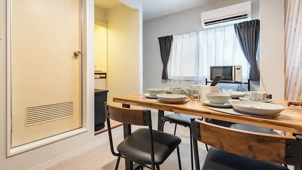 A dining area with a wooden table set for four, a black chair, and a kitchen in the background. - Wagyu House (Osaka)