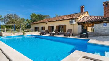 A swimming pool with lounge chairs, a house with a tiled roof, and a stone wall.