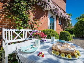 A table set outdoors with a cake, a white bench, and a brick building in the background.