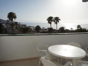 A balcony with a round table and chairs, overlooking a coastal area with palm trees and buildings. - Apartment Tenerife Near Sea & Pool (Santiago del Teide)