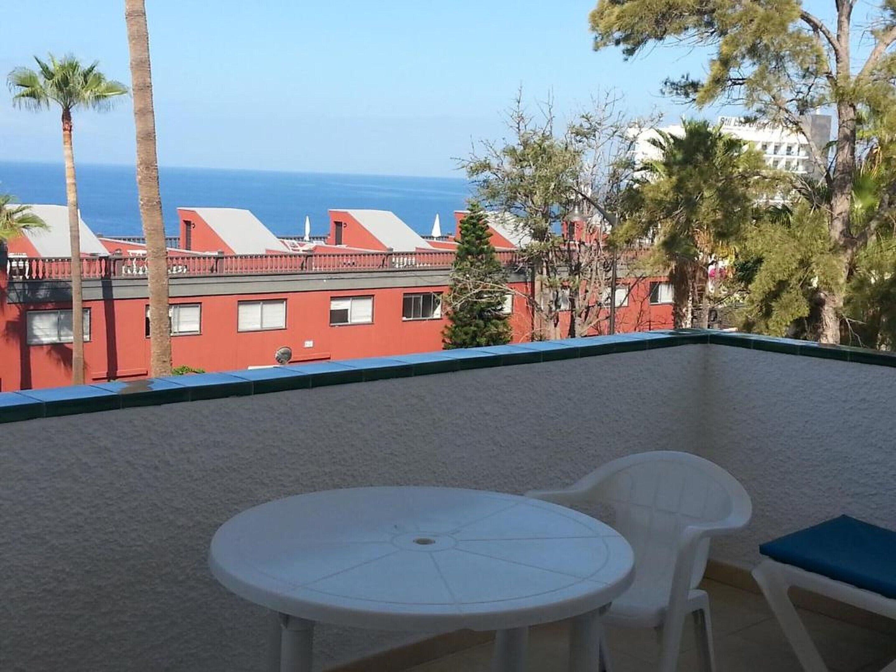 Ein Balkon mit weißem Tisch und zwei Stühlen, mit Blick auf eine Küstenlandschaft mit roten Gebäuden und Palmen.