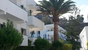 A modern building with white exterior walls, multiple balconies, and lush greenery including palm trees and shrubs. - Apartment Tenerife Near Sea & Pool (Santiago del Teide)