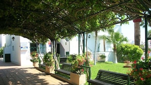 A covered walkway with benches and planters, leading to a building with a white facade and a signboard. - Apartment Tenerife Near Sea & Pool (Santiago del Teide)