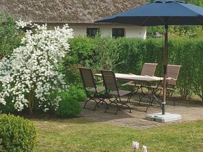 Een terras buiten met een tafel en stoelen, een gebouw met rieten dak en een bloeiende struik.