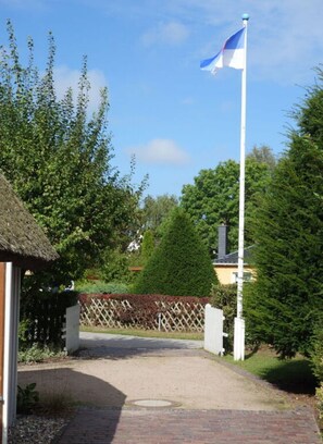 Enter a serene garden surrounded by lush trees and shrubs, with a traditional thatched-roof structure and a charming picket fence.