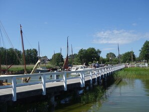 Cross a tranquil bridge to the riverbank, where sailboats await and lush greenery surrounds.
