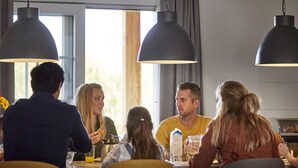 Eine Gruppe von Menschen speist an einem Tisch, beleuchtet von Pendelleuchten, mit großen Fenstern und Blick ins Freie.