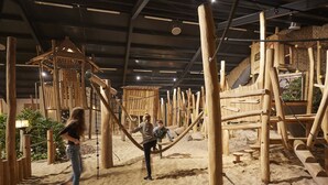 Eine hölzerne Spielkonstruktion in einem sandigen Innenbereich mit Kletternetzen und Seilen.