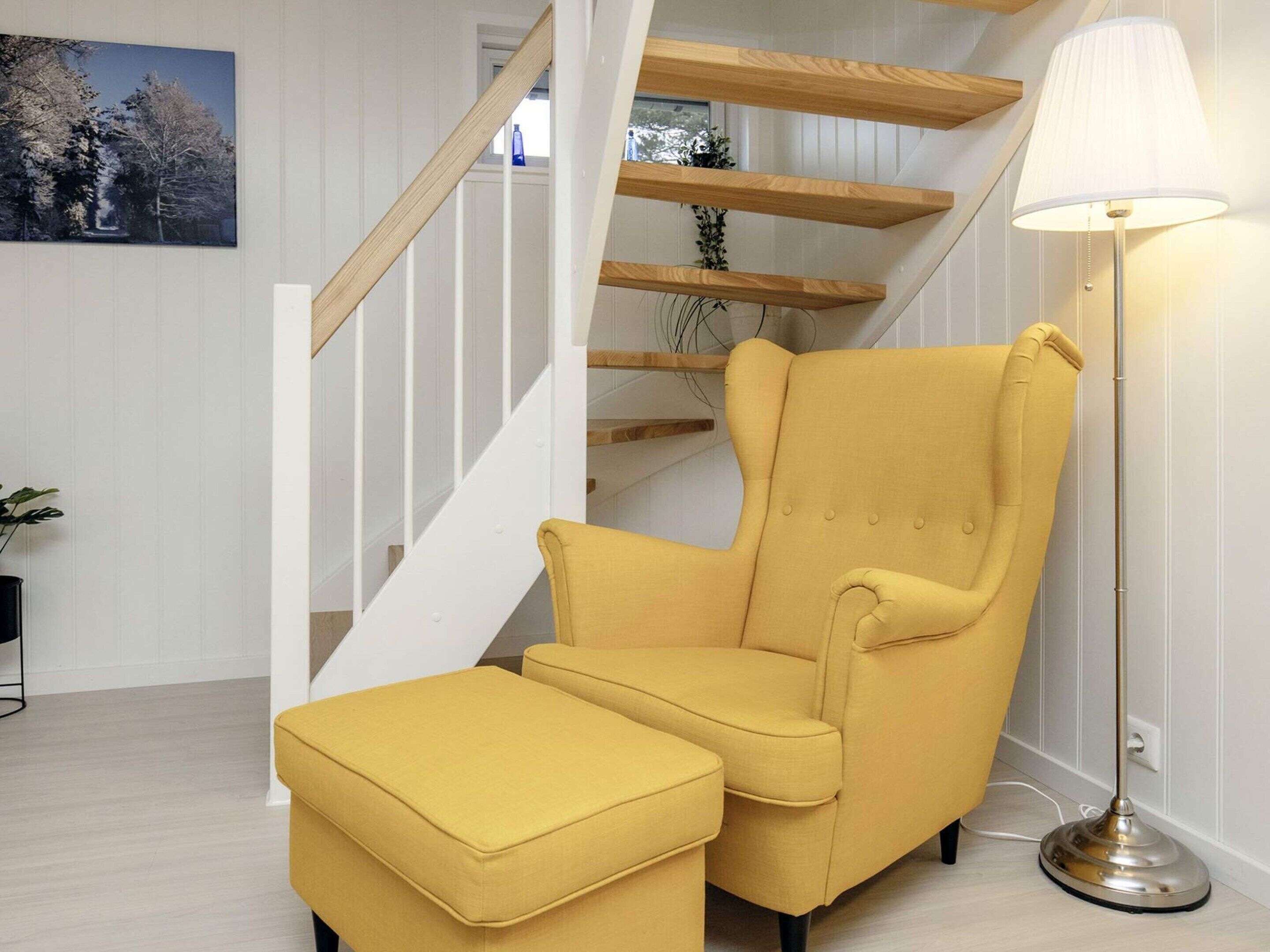 Ett modernt vardagsrum med en gul fåtölj och ett tillhörande fotstöd, en vit trappa och en golvlampa.