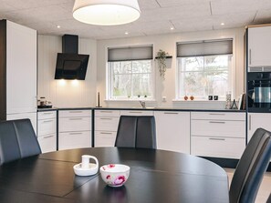 Une cuisine moderne avec des meubles blancs, une table à manger avec des chaises, un téléviseur fixé au-dessus du comptoir et une fenêtre avec des stores.
