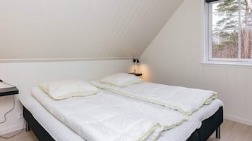 Un dormitorio con techo inclinado, una cama con ropa de cama blanca, una mesita de noche con lámpara y una ventana con vistas a árboles.