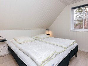 Une chambre avec un plafond en pente, un lit avec une literie blanche, une table de chevet avec une lampe et une fenêtre donnant sur des arbres.