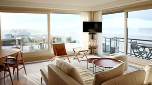 Ein geräumiges Wohnzimmer mit einer großen Couch, einem Couchtisch und einem an der Wand montierten Fernseher. Ein Balkon mit Glasgeländer und Gartenmöbeln ist ebenfalls vorhanden.