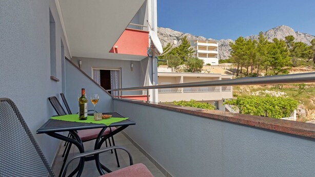 A balcony with a table set for two, a bottle of wine, and a glass. The balcony overlooks a mountainous landscape with buildings in the background. - Apartments Villa Juric - One Bedroom Apartment (Baska Voda)