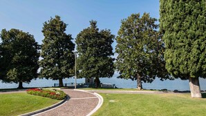 A paved pathway curving through a park with large trees and a body of water in the background. - Belvilla by OYO Comfortable Apartment With Pool (Garda)