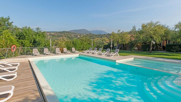 A swimming pool with lounge chairs and a view of a green landscape with trees and hills. - Belvilla by OYO Comfortable Apartment With Pool (Garda)