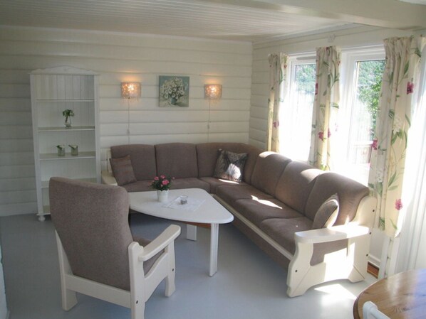 A cozy living room with a brown sectional sofa, a white coffee table, and a white armchair. There is a white cabinet with shelves and a small plant on the left.