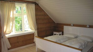 Una camera da letto con soffitto spiovente in legno, un letto singolo con biancheria bianca, un comodino e una finestra con tende.