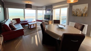 Een ruime woonkamer met een groot raam met uitzicht op het strand, een eethoek en een televisie.