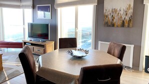 Een moderne woonkamer met een eettafel, stoelen, een televisietoestel en grote ramen met uitzicht op de stad.