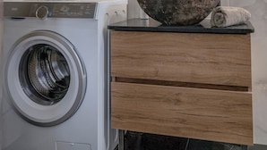 A modern bathroom with a built-in washing machine, a wooden vanity, and a large mirror with a unique curved frame. - Calliope's Rhodes City Comfort (Rhodes)