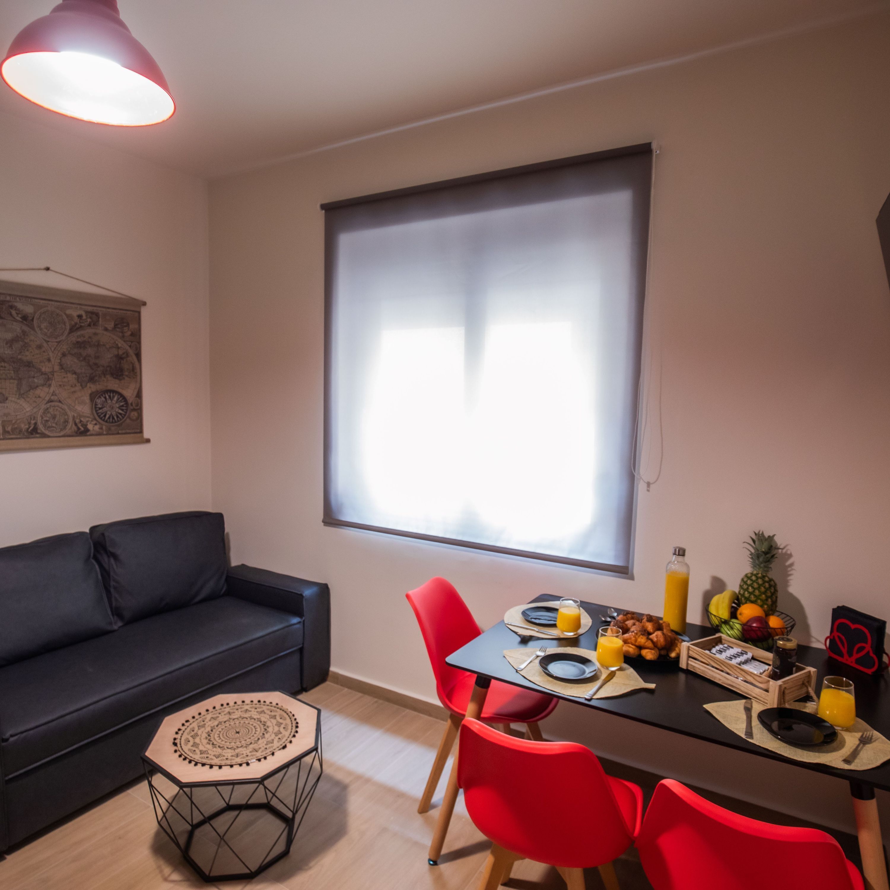Un comedor moderno con una mesa preparada para dos, sillas rojas, un sofá negro, una mesita hexagonal y un ventanal con persianas.