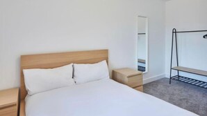 Una camera da letto con un letto in legno, un comodino, una piccola cassettiera in legno, una scaffalatura metallica e uno specchio appeso al muro.