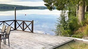 A wooden deck with chairs, a lifebuoy, and a railing overlooking a calm lake with trees. - 4 Star Holiday Home in Tronodal (Trönödal)
