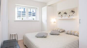 Un dormitorio con cama, almohadas, mesita de noche, silla, ventana y estante con adornos.