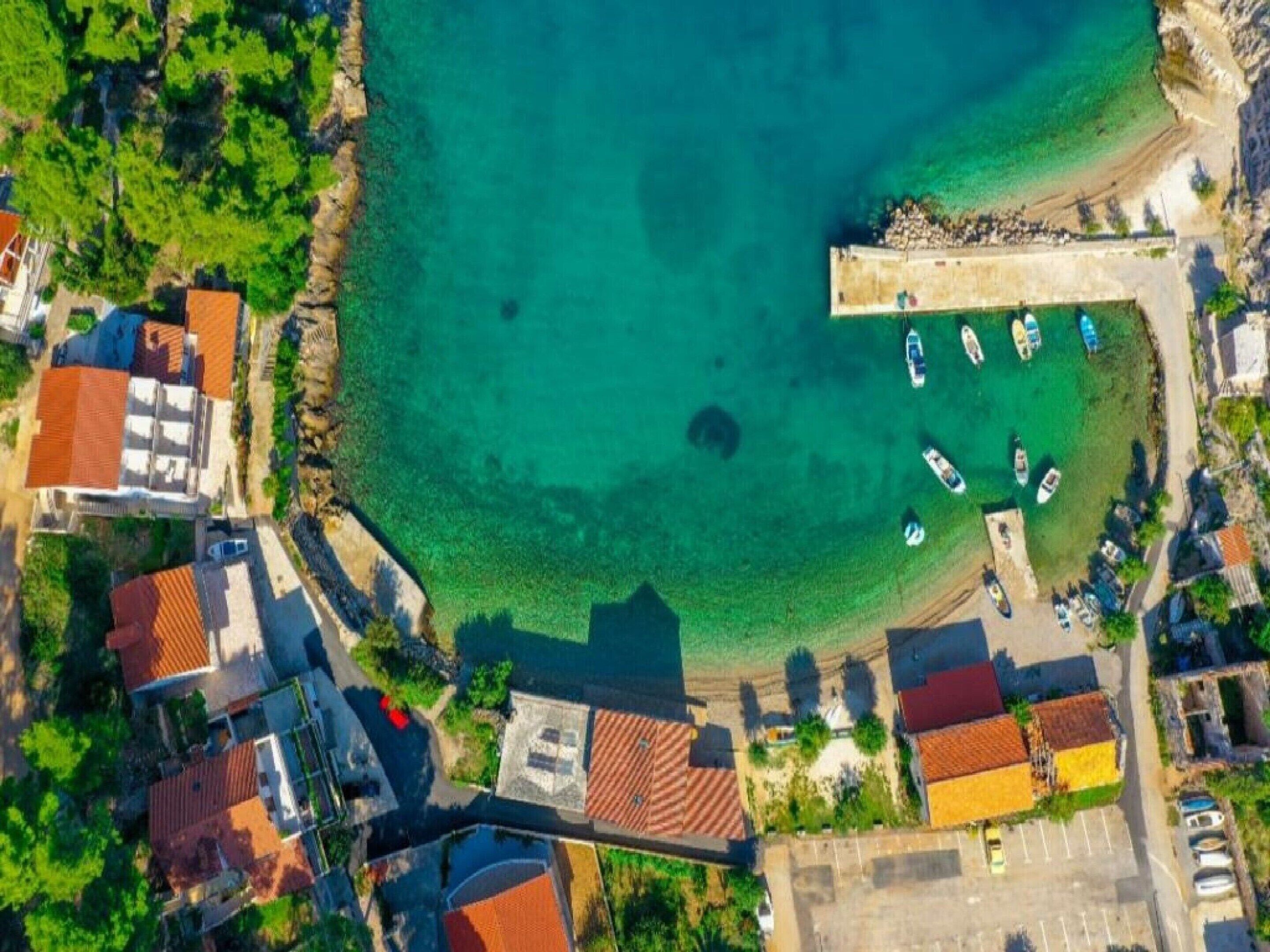 An aerial view of a coastal area with clear turquoise waters, a marina with several boats, and buildings with red-tiled roofs.