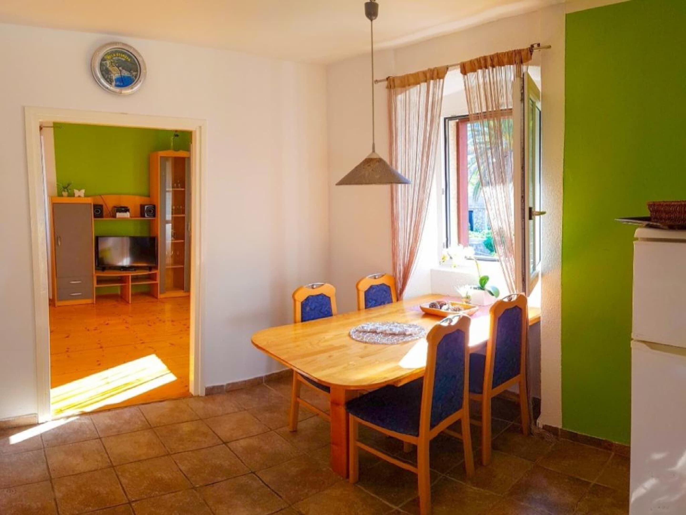 Un coin repas avec une table en bois et des chaises bleues, une cuisine en arrière-plan et un mur vert.