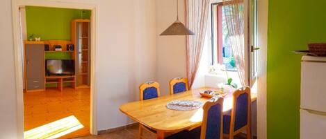 A dining area with a wooden table and blue chairs, a kitchen in the background, and a green wall.