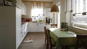 A compact kitchen with white cabinets, a dining table with a green tablecloth, wooden chairs, and a hanging pendant light. - Holiday Home Sonnentau (Burg (Spreewald))